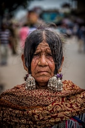 An elderly woman with a solemn expression, adorned in traditional attire. Her hair is parted in the middle, and she wears large, ornate earrings and a richly patterned shawl. The background is slightly blurred, with people walking in a street-like setting.