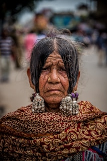 An elderly woman with a solemn expression, adorned in traditional attire. Her hair is parted in the middle, and she wears large, ornate earrings and a richly patterned shawl. The background is slightly blurred, with people walking in a street-like setting.
