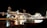 An industrial facility is illuminated at night, with numerous lights highlighting its intricate structure. The scene includes various industrial buildings, pipes, and machinery with a calm body of water reflecting the lights. The water is bordered by rocks and some low vegetation in the foreground.