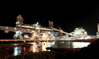 An industrial facility is illuminated at night, with numerous lights highlighting its intricate structure. The scene includes various industrial buildings, pipes, and machinery with a calm body of water reflecting the lights. The water is bordered by rocks and some low vegetation in the foreground.