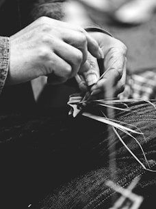 A close-up black and white photo of hands weaving threads, suggesting skilled craftsmanship or an artisanal process. The hands are manipulating thin, delicate strands, indicative of intricate handiwork.