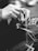 A close-up black and white photo of hands weaving threads, suggesting skilled craftsmanship or an artisanal process. The hands are manipulating thin, delicate strands, indicative of intricate handiwork.