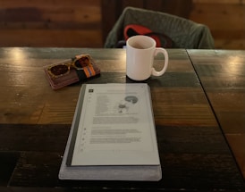 A tablet displaying a document is placed on a wooden table alongside a white mug, a pair of sunglasses, and a wallet. There is a chair with a quilted jacket hanging on it in the background.
