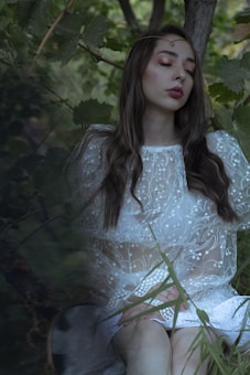 A woman in a delicate, sheer white lace dress sits surrounded by lush green foliage. Her long hair flows naturally, and she has an expression of serene tranquility with her eyes closed. The scene conveys a sense of peaceful solitude within nature.