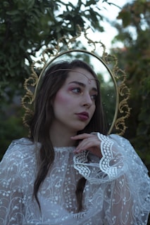 A woman with long, dark hair wearing a delicate lace garment that features intricate floral embroidery. She is positioned against a backdrop of lush green foliage and wears a gold metallic headpiece resembling a halo. Her makeup includes soft pink blush and eyeshadow. She gazes thoughtfully into the distance with her hand gently placed on her chin.