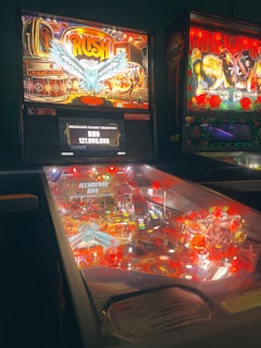 The image features a pinball machine with a vibrant and colorful design. The backboard displays graphics of a stylized fantasy scene with an owl and the word 'Rush' in bold letters. The pinball table is brightly lit with various flashing lights and bumpers, indicating an active game in progress. To the right, another pinball machine featuring the theme of 'Guns N' Roses' is partially visible.