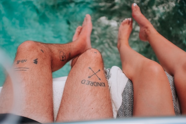 Two tattooed legs dangle over a body of clear turquoise water. The leg on the left features multiple tattoos, including the word 'KINDRED' and small symbols. Both legs rest on a white towel with a black geometric pattern.