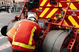 A firefighter in protective gear is operating a fire truck equipped with hoses and various control panels. The truck is marked with bold red and yellow stripes, and there are visible text labels indicating it's part of the Paris fire department.