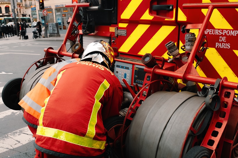 A firefighter in protective gear is operating a fire truck equipped with hoses and various control panels. The truck is marked with bold red and yellow stripes, and there are visible text labels indicating it's part of the Paris fire department.