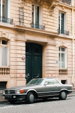 A classic silver Mercedes-Benz convertible is parked on a cobblestone street in front of a historic European-style building with ornate architectural details and large green double doors.