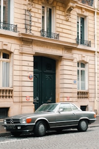 A classic silver Mercedes-Benz convertible is parked on a cobblestone street in front of a historic European-style building with ornate architectural details and large green double doors.