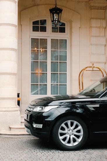 A black luxury SUV is parked in front of a grand, ivory-colored building with large windows and an ornate hanging lantern. The building's entrance is elegant, featuring a light stone facade with tall arched windows and a decorative gold luggage cart nearby.