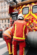 Two firefighters wearing red protective uniforms with reflective stripes are standing next to a fire truck with bright red and yellow stripes. The fire truck is parked on a city street, and the firefighters appear to be tending to equipment on the truck. Nearby, a building with decorative balconies and windows is visible in the background.