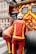 Two firefighters wearing red protective uniforms with reflective stripes are standing next to a fire truck with bright red and yellow stripes. The fire truck is parked on a city street, and the firefighters appear to be tending to equipment on the truck. Nearby, a building with decorative balconies and windows is visible in the background.