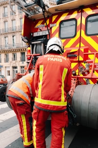 Two firefighters wearing red protective uniforms with reflective stripes are standing next to a fire truck with bright red and yellow stripes. The fire truck is parked on a city street, and the firefighters appear to be tending to equipment on the truck. Nearby, a building with decorative balconies and windows is visible in the background.