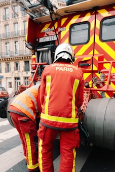 Two firefighters wearing red protective uniforms with reflective stripes are standing next to a fire truck with bright red and yellow stripes. The fire truck is parked on a city street, and the firefighters appear to be tending to equipment on the truck. Nearby, a building with decorative balconies and windows is visible in the background.