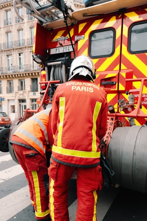 Two firefighters wearing red protective uniforms with reflective stripes are standing next to a fire truck with bright red and yellow stripes. The fire truck is parked on a city street, and the firefighters appear to be tending to equipment on the truck. Nearby, a building with decorative balconies and windows is visible in the background.