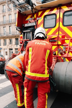 Two firefighters wearing red protective uniforms with reflective stripes are standing next to a fire truck with bright red and yellow stripes. The fire truck is parked on a city street, and the firefighters appear to be tending to equipment on the truck. Nearby, a building with decorative balconies and windows is visible in the background.