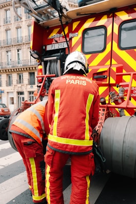 Two firefighters wearing red protective uniforms with reflective stripes are standing next to a fire truck with bright red and yellow stripes. The fire truck is parked on a city street, and the firefighters appear to be tending to equipment on the truck. Nearby, a building with decorative balconies and windows is visible in the background.