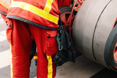 A person is wearing a red uniform with bright yellow reflective stripes, standing next to a large industrial hose reel. Black gloves are tucked into the side of the uniform.