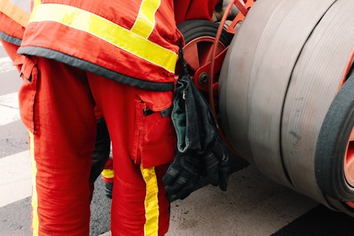 A person is wearing a red uniform with bright yellow reflective stripes, standing next to a large industrial hose reel. Black gloves are tucked into the side of the uniform.