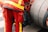 A person is wearing a red uniform with bright yellow reflective stripes, standing next to a large industrial hose reel. Black gloves are tucked into the side of the uniform.