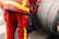 A person is wearing a red uniform with bright yellow reflective stripes, standing next to a large industrial hose reel. Black gloves are tucked into the side of the uniform.