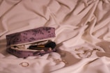A floral-patterned makeup bag rests on soft white fabric. The bag is unzipped to reveal a makeup brush inside. Nearby, a gold chain, rings, and earrings are scattered on the fabric, creating an elegant and organized display.
