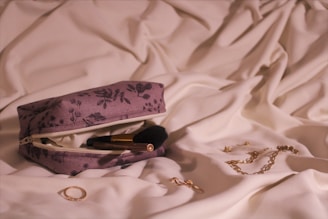 A floral-patterned makeup bag rests on soft white fabric. The bag is unzipped to reveal a makeup brush inside. Nearby, a gold chain, rings, and earrings are scattered on the fabric, creating an elegant and organized display.