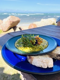 A vibrant meal with a creamy, herb-infused dish on a blue plate, accompanied by slices of rustic bread. The dish is garnished with fresh greens and possibly salmon, with an expansive ocean view and rocky shoreline in the background.