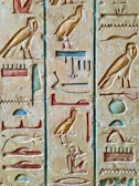 The image features detailed ancient Egyptian hieroglyphs carved on a stone surface. The carvings are filled with colors such as blue, red, and gold, representing various symbols like birds, seated figures, and abstract shapes. The artwork showcases the intricate craftsmanship and historical significance of ancient Egyptian writing.