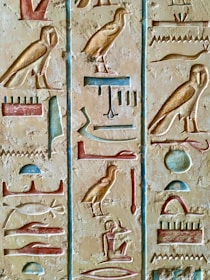 The image features detailed ancient Egyptian hieroglyphs carved on a stone surface. The carvings are filled with colors such as blue, red, and gold, representing various symbols like birds, seated figures, and abstract shapes. The artwork showcases the intricate craftsmanship and historical significance of ancient Egyptian writing.