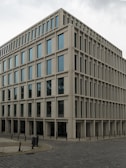 A modern, rectangular office building with multiple floors and large windows. The building has a uniform, grid-like structure and is situated on a cobblestone corner in an urban area.