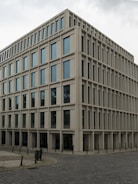 A modern, rectangular office building with multiple floors and large windows. The building has a uniform, grid-like structure and is situated on a cobblestone corner in an urban area.