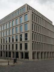 A modern, rectangular office building with multiple floors and large windows. The building has a uniform, grid-like structure and is situated on a cobblestone corner in an urban area.