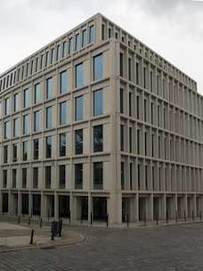 A modern, rectangular office building with multiple floors and large windows. The building has a uniform, grid-like structure and is situated on a cobblestone corner in an urban area.