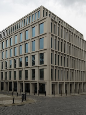 A modern, rectangular office building with multiple floors and large windows. The building has a uniform, grid-like structure and is situated on a cobblestone corner in an urban area.