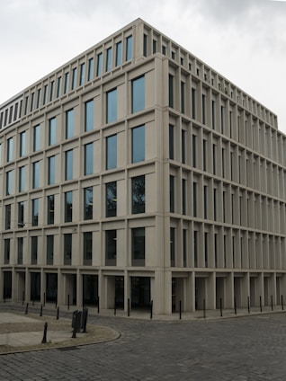A modern, rectangular office building with multiple floors and large windows. The building has a uniform, grid-like structure and is situated on a cobblestone corner in an urban area.