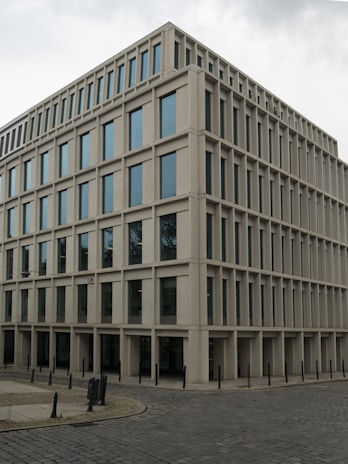 A modern, rectangular office building with multiple floors and large windows. The building has a uniform, grid-like structure and is situated on a cobblestone corner in an urban area.