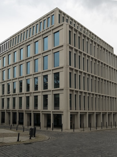 A modern, rectangular office building with multiple floors and large windows. The building has a uniform, grid-like structure and is situated on a cobblestone corner in an urban area.