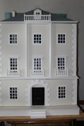 A detailed view of a three-story model building with a classical architectural design, featuring multiple white framed windows and a prominent black central door. The facade includes decorative columns and railings, and the roof is a muted gray.