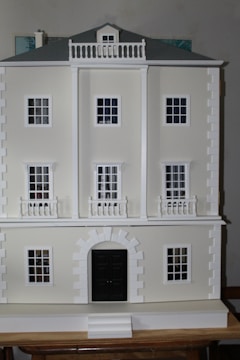 A detailed view of a three-story model building with a classical architectural design, featuring multiple white framed windows and a prominent black central door. The facade includes decorative columns and railings, and the roof is a muted gray.