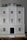 A detailed view of a three-story model building with a classical architectural design, featuring multiple white framed windows and a prominent black central door. The facade includes decorative columns and railings, and the roof is a muted gray.