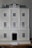 A detailed view of a three-story model building with a classical architectural design, featuring multiple white framed windows and a prominent black central door. The facade includes decorative columns and railings, and the roof is a muted gray.