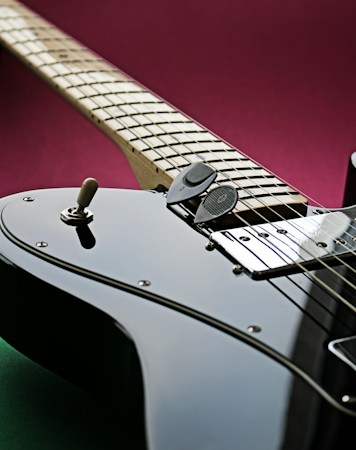 A close-up view of an electric guitar displaying intricate details of its strings, pickups, and controls. The guitar is angled diagonally, with a focus on the fretboard and shiny black body. A deep magenta background contrasts with the instrument's sleek appearance.