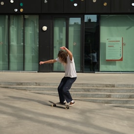 A person is skateboarding on a concrete surface near steps, next to a modern building with large glass windows and a dark facade adorned with circular decorations. The person is wearing a white shirt and dark pants, and they are positioned in a dynamic pose, suggesting movement.