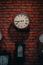Several antique clocks are mounted on a rustic, exposed brick wall. The central clock features Roman numerals and inscriptions on its face, surrounded by a wooden frame. Additional smaller clocks, each with distinct designs, contribute to a vintage ambiance.