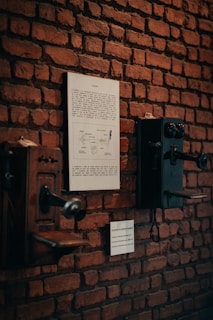 Vintage telephones mounted on a rustic brick wall, accompanied by informational plaques providing historical context. The arrangement creates a museum-like display with an antique feel.