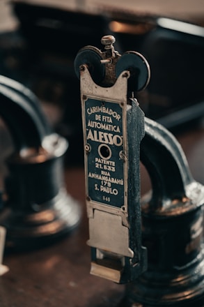 A vintage metal device with a label in Portuguese indicating it is an automatic ribbon stamper patented in São Paulo. The intricate machinery features wheels and knobs, emphasizing its industrial and historical design.