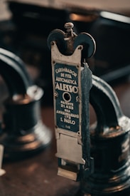 A vintage metal device with a label in Portuguese indicating it is an automatic ribbon stamper patented in S&atilde;o Paulo. The intricate machinery features wheels and knobs, emphasizing its industrial and historical design.